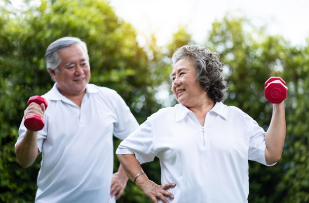Smiling seniors in white shirts exercise outdoors with red dumbbells, enjoying a bright, green background.